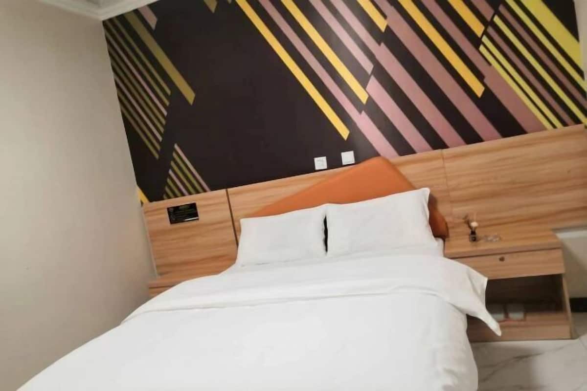 Un lit bien fait, avec une literie blanche et un oreiller orange, une tête de lit en bois et une petite table de chevet avec une lampe.
