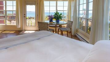 A bedroom with a large bed, a dining table with chairs, and a view of the sea through multiple windows.