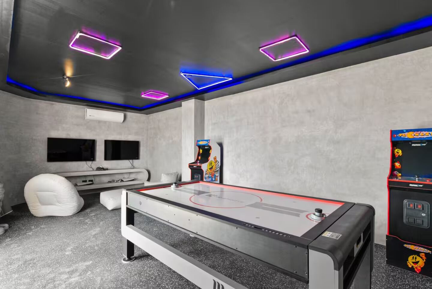 A room with a pool table, two flat-screen TVs, a white chair, and arcade games.
