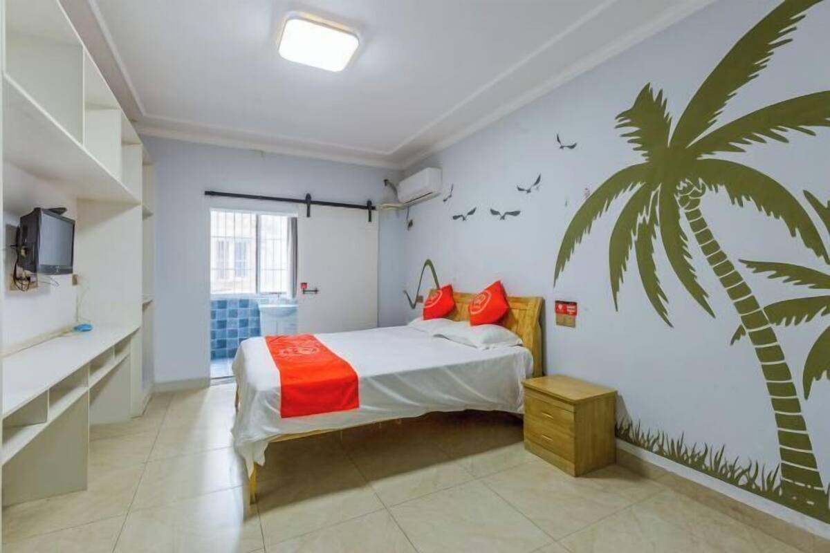 Un dormitorio con una cama, una mesita de noche, un televisor sobre una estantería y una pared con un mural de palmeras.