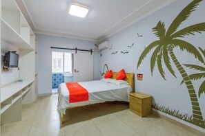 Une chambre à coucher avec un lit, une table de chevet, un téléviseur posé sur une étagère et un mur orné d’une fresque représentant un palmier.