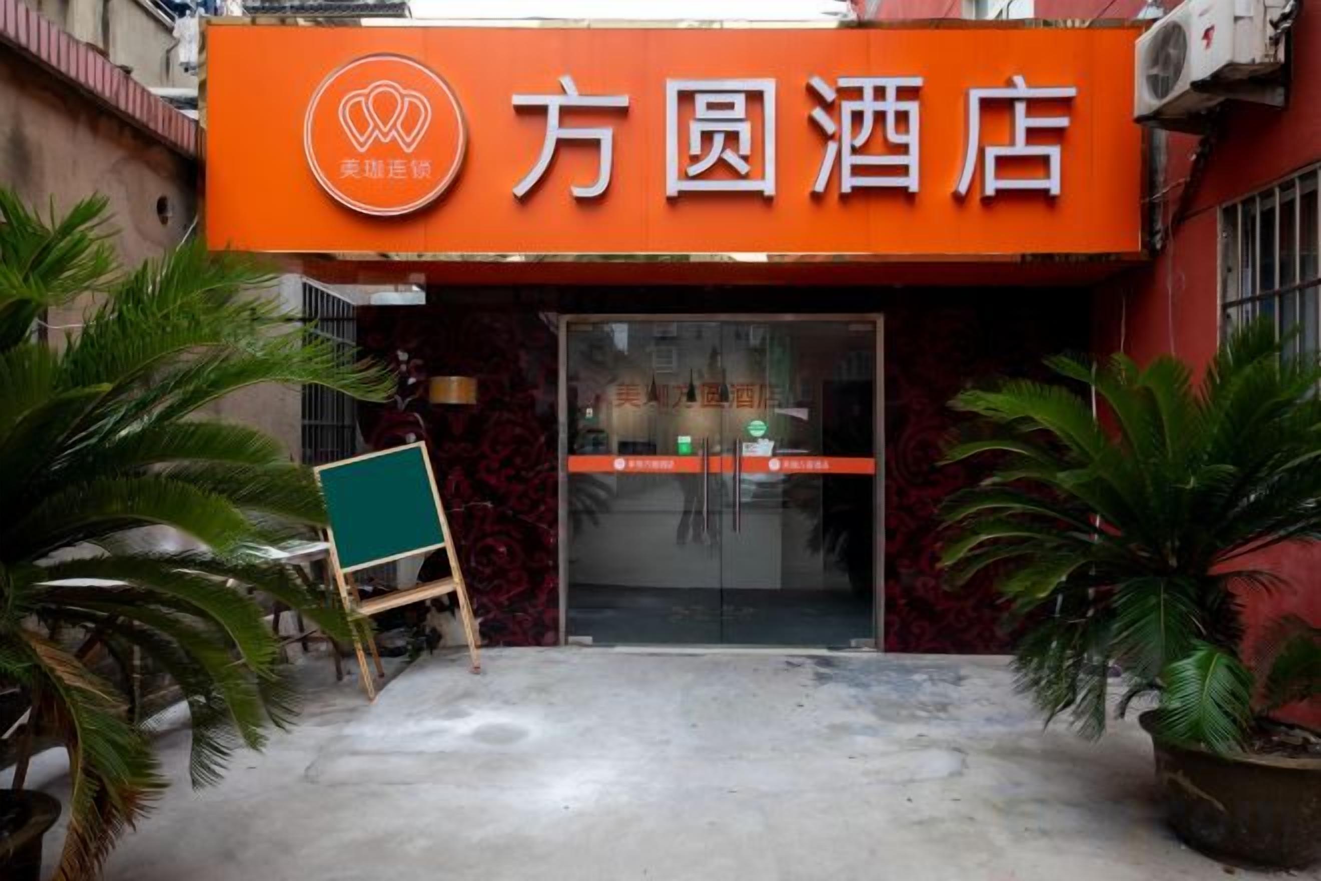 Entrada de hotel com placa laranja chamativa, porta de vidro e plantas em vasos.