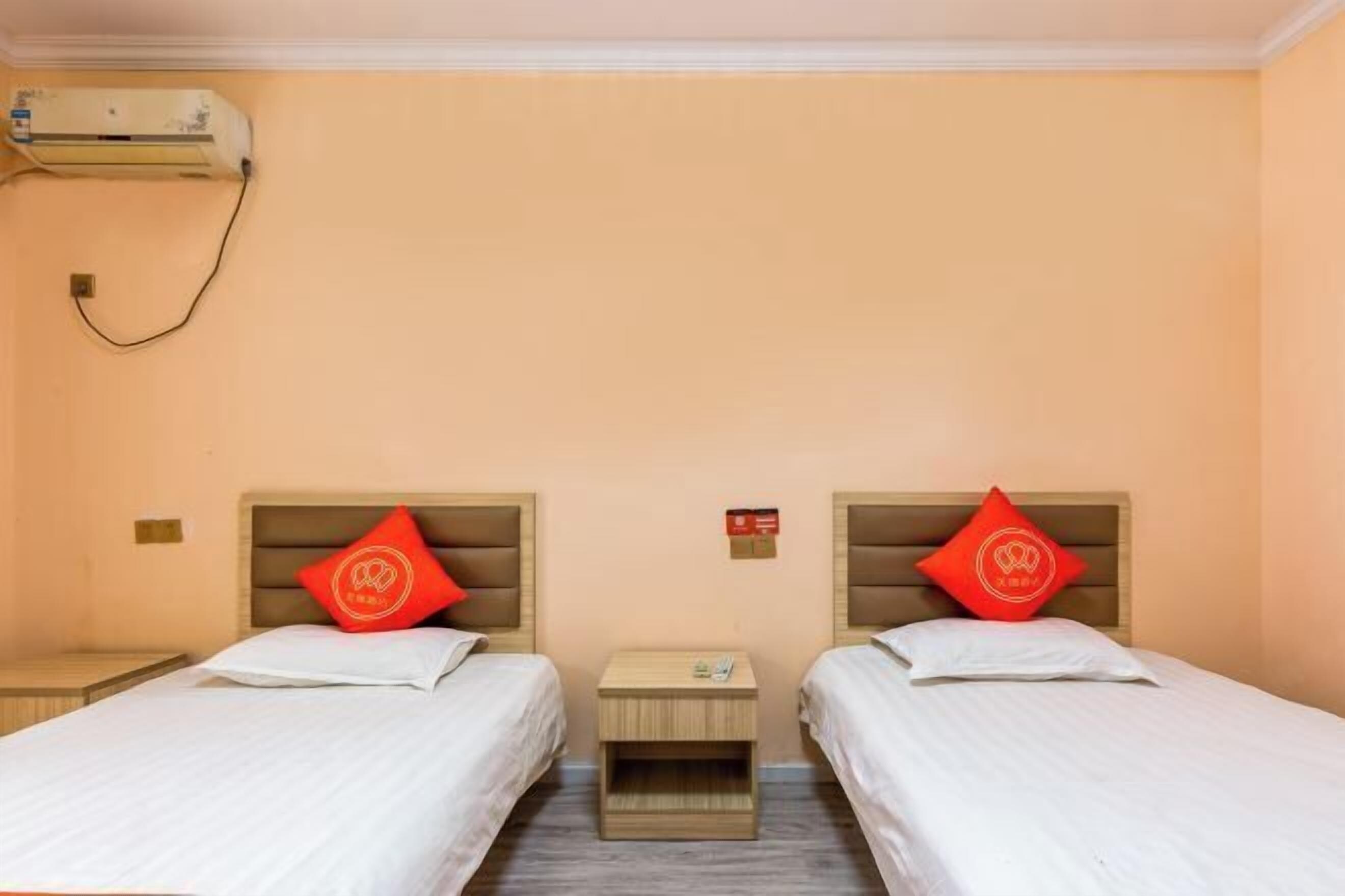 Habitación de hotel con dos camas individuales, cada una con cabecero de madera y almohada roja. Un pequeño mesita de noche de madera entre las camas contiene un teléfono y una pequeña mesa. Un equipo de aire acondicionado está montado en la pared sobre las camas.