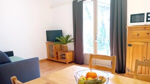 A dining area with a wooden table, chairs, a fruit bowl, a television on a wooden cabinet, and a window with curtains. - Mazet Provençal air-conditioned swimming pool parking 5 pers (Gaujac)
