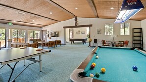 A pool table with billiard balls, a ping pong table, and a billiards table in a room with wooden ceiling beams, large windows, and a Coors Light sign.