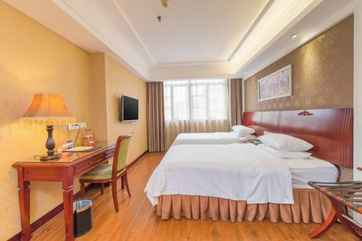 A hotel room with two beds, a desk, a chair, a TV, and a window with curtains.