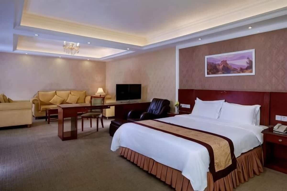 A hotel room with a bed, a sofa, a desk with a TV, and a painting on the wall.