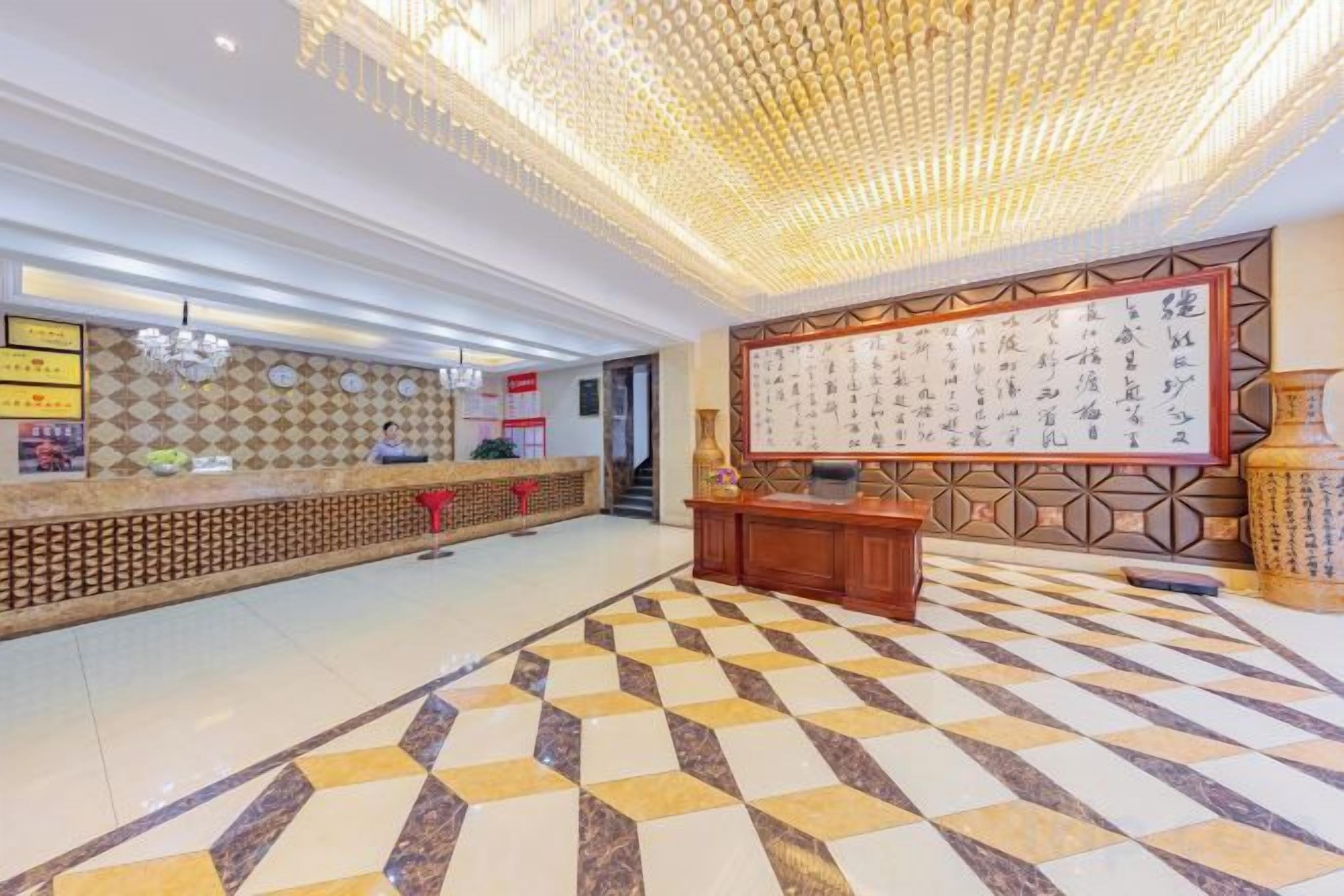A hotel lobby with a reception desk, a large wall with calligraphy, and a patterned floor.