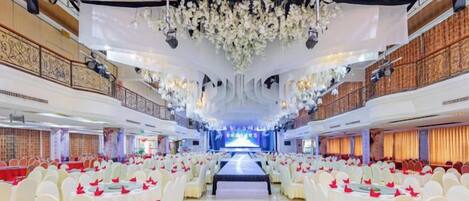 A banquet hall with rows of white chairs, a stage with a truss system, and a balcony on one side.
