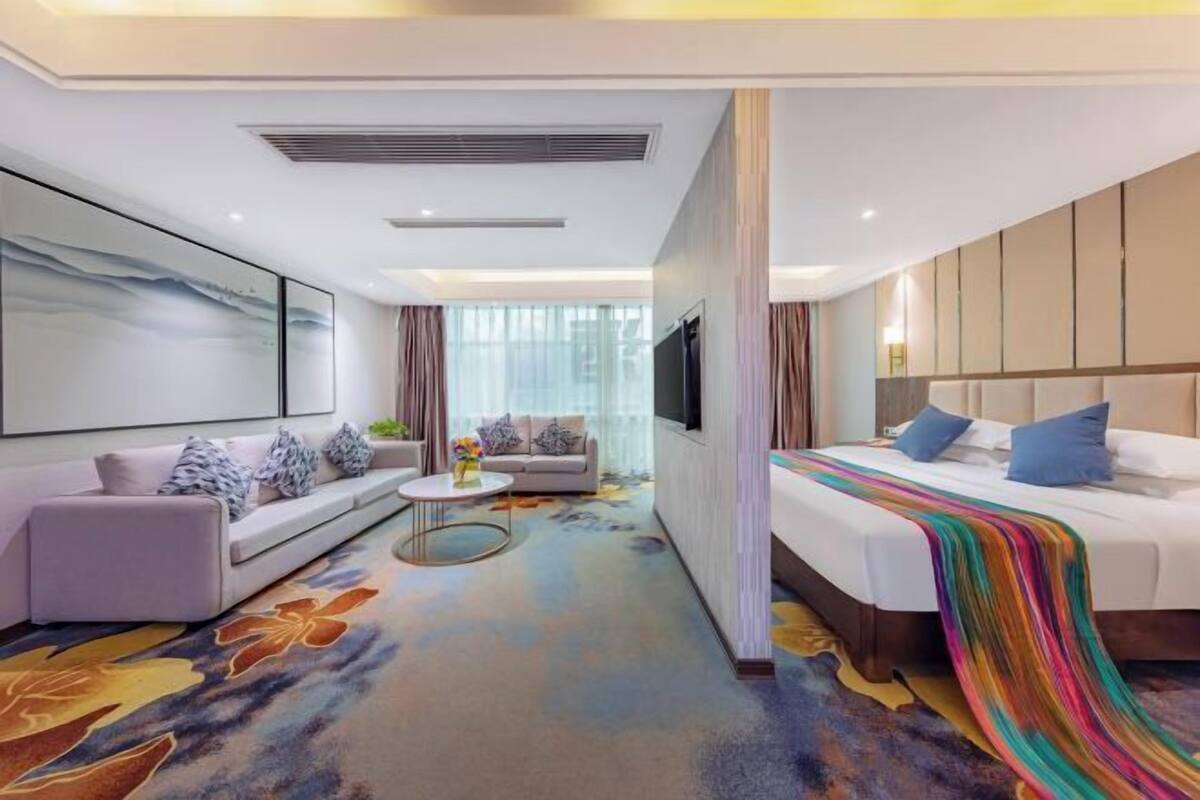 A modern hotel room with a sofa, a bed with a colorful blanket, a TV, and a large window with curtains.