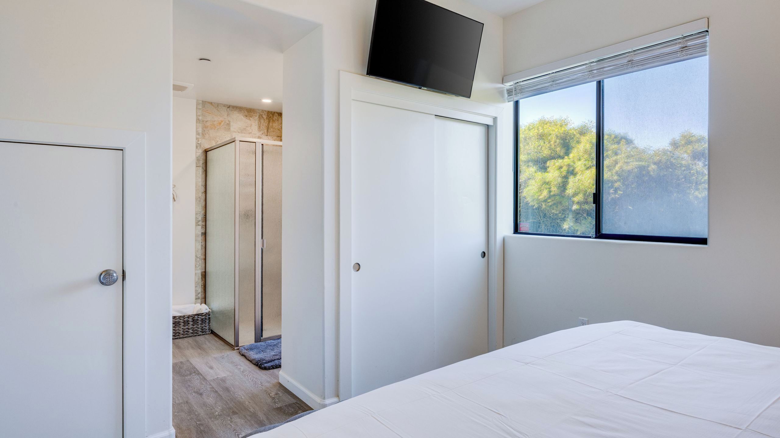 Un dormitorio con cama, ventana con vistas a árboles, zona de ducha y puerta blanca.