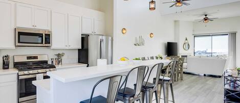 Una cocina moderna con gabinetes blancos, electrodomésticos de acero inoxidable, una isla central con taburetes altos e iluminación colgante.