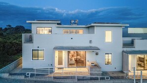 A modern two-story house with a flat roof, large windows, and a glass door leading to an interior with a sofa and coffee table. - Beachfront 2BR w/ Deck, Views & Direct Sand Access Strand Way A (Oceano)