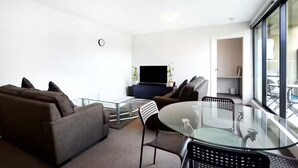 A modern living room with a glass coffee table, two sofas, a TV, and a large window.