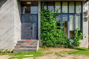 Ein modernes Gebäude mit einer Betonfassade, einer Holztreppe, die zu einer dunklen Tür führt, und einem Busch vor der Fassade.