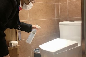 A person holding a spray bottle in a bathroom with a toilet and a trash bin.