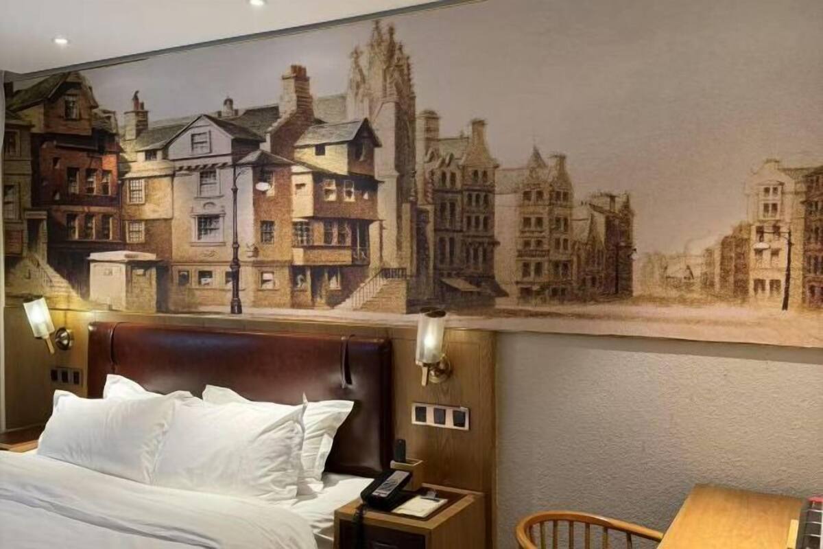 Hotelkamer met een groot muurschildering van een Europese stad, een houten hoofdeinde, een nachtkastje met een lamp en een stoel.