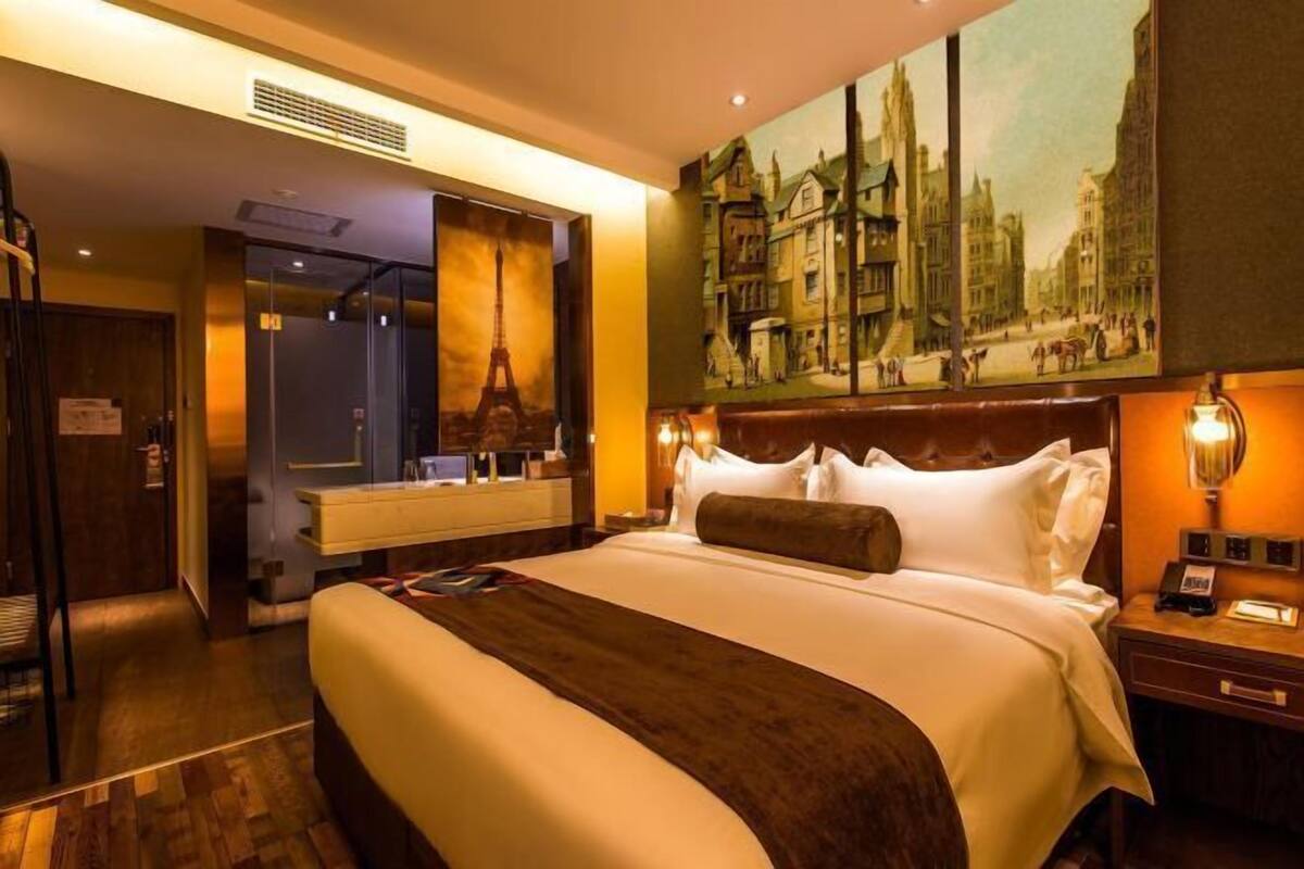 Hotelkamer met een groot bed, een schilderij van een stadsgezicht, een houten hoofdbord en een nachtkastje met een lamp.