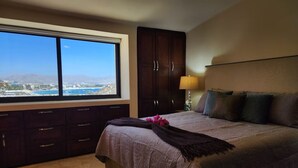 A bedroom with a large window, a bed with multiple pillows, a nightstand, and a lamp.