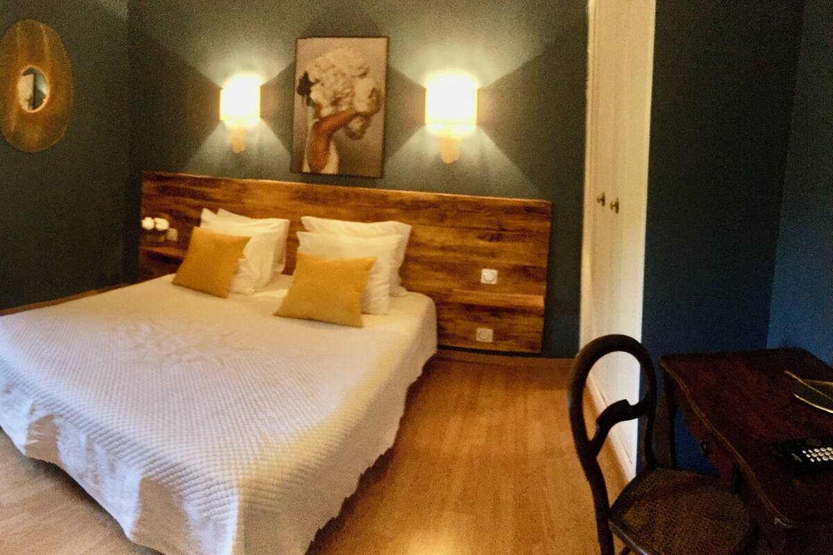 Une chambre avec un grand lit, une tête de lit en bois, deux lampes fixées au mur, une chaise et une petite table avec une télécommande.