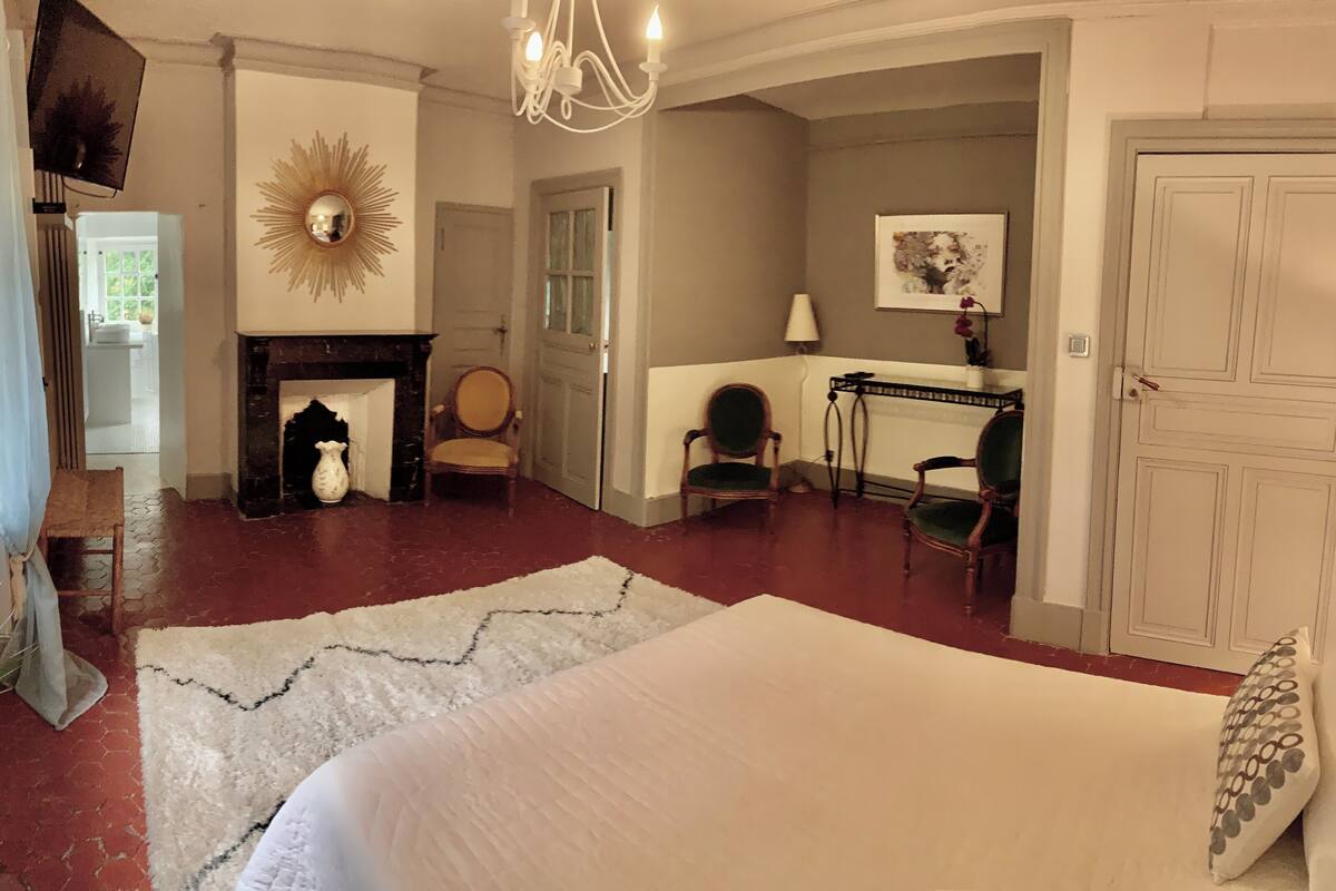 Une chambre à coucher avec un grand lit, une cheminée, deux fauteuils, une table et une porte.