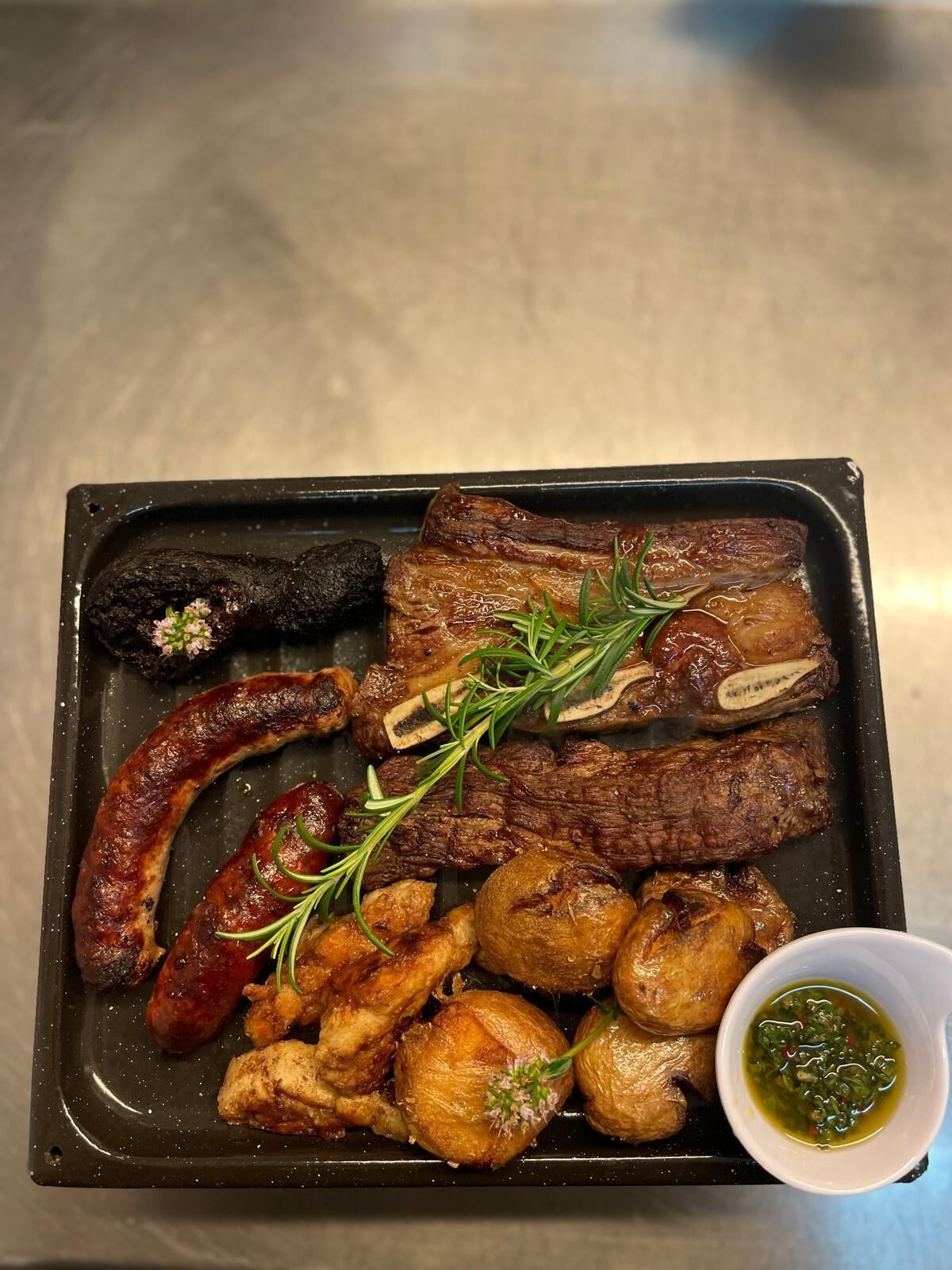 Una bandeja de carnes y salchichas a la parrilla, acompañada de patatas asadas y un recipiente pequeño con salsa.