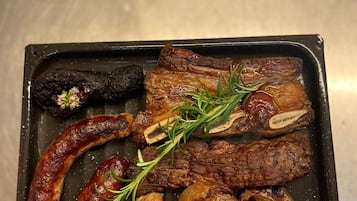 Una bandeja de carnes y salchichas a la parrilla, acompañada de patatas asadas y un recipiente pequeño con salsa.