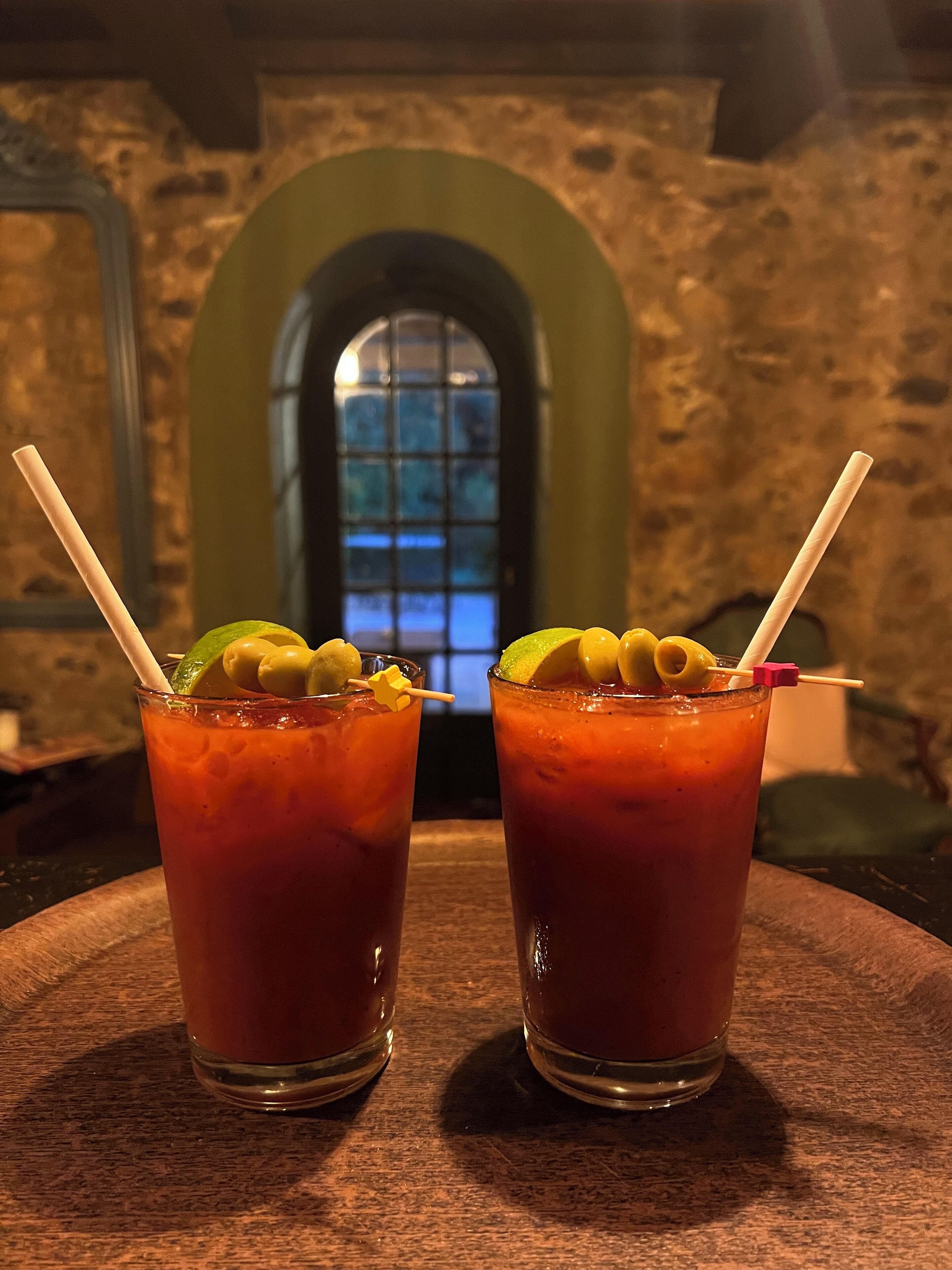 Two glasses of a red beverage with lime and olives, placed on a wooden table with a stone wall and an arched window in the background.