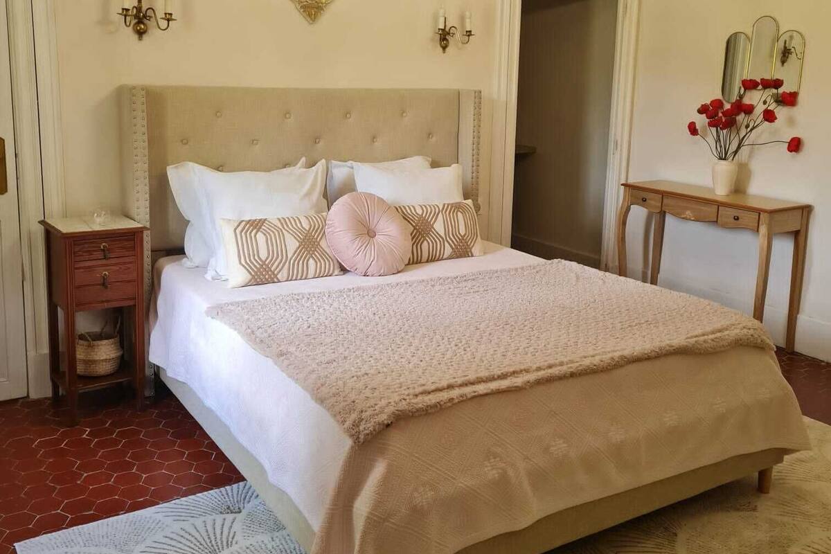 Une chambre à coucher avec un grand lit, un lustre, un élément décoratif mural et une petite table sur laquelle est posé un vase.