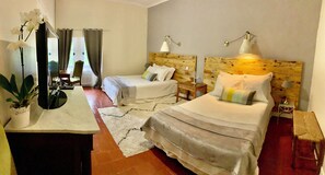 Habitación de hotel con dos camas, cabecera de madera, mesita de noche, lámpara, espejo, planta en maceta y vista al exterior a través de una ventana.