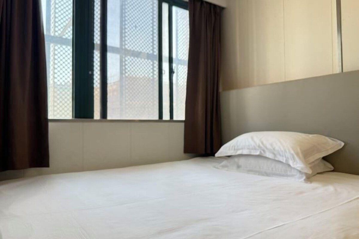 Una cama individual con ropa blanca, una ventana con mosquitero y cortinas marrones.