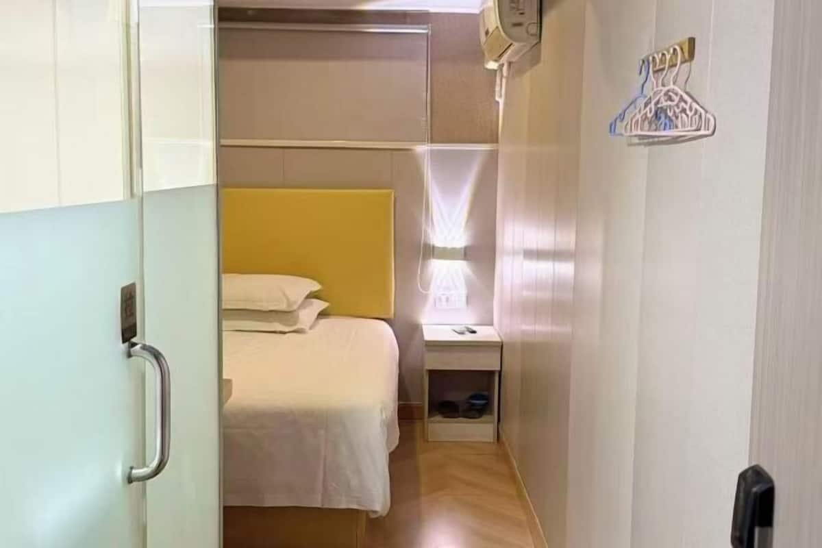 Une chambre d’hôtel étroite, comprenant un lit, une table de chevet et un porte-manteau mural.