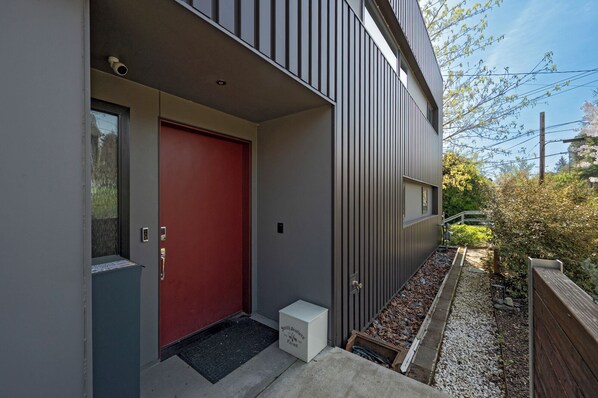 A modern building with a red door, a small porch area, and a garden with gravel paths. - Modern 3BR Retreat w/ Rooftop Hot Tub, Firepit & City Views (Seattle)