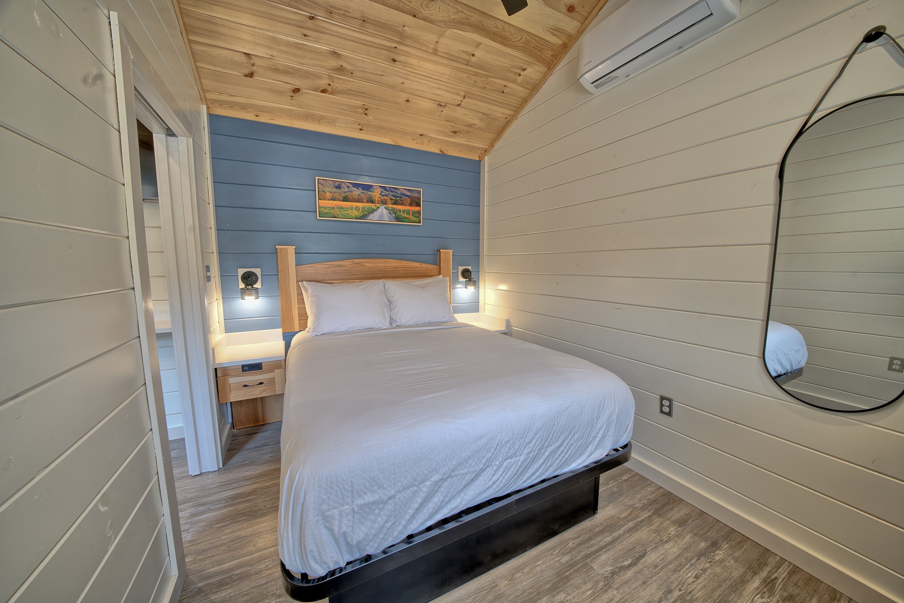 A bedroom with a wooden ceiling, a bed with white bedding, a nightstand, a wall-mounted mirror, and a painting on the wall.