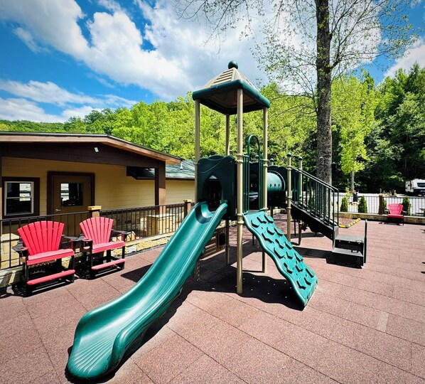 A playground with a slide, climbing structures, and red Adirondack chairs. - Dudley Creek RV Resort (Gatlinburg)