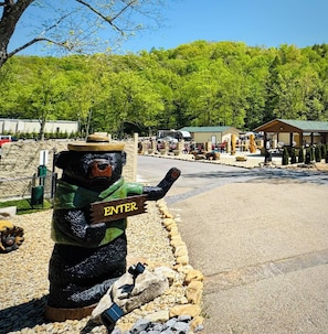 A bear statue with an 'ENTER' sign, surrounded by pebbles and rocks, with a forested background and a building in the distance.