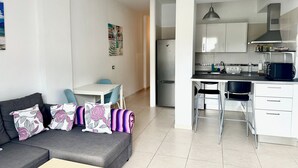 Um apartamento moderno com sala de estar, que inclui um sofá cinza, uma mesa de jantar com cadeiras azuis e uma cozinha com armários brancos, micro-ondas preto e geladeira de aço inoxidável.