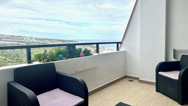 Uma varanda com duas cadeiras de vime, vista para o mar e paisagem urbana ao fundo.