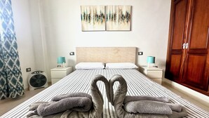 Quarto de hotel com cama listrada, duas toalhas dispostas em formato de coração, cabeceira de madeira e um guarda-roupa grande.