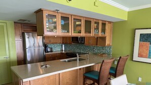 A kitchen with wooden cabinets, a stainless steel refrigerator, a microwave, and a glass island with chairs.