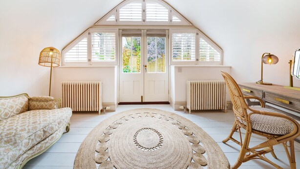 Una habitación con techo alto, un ventanal con contraventanas, un radiador, un sofá, una silla y una alfombra redonda.
