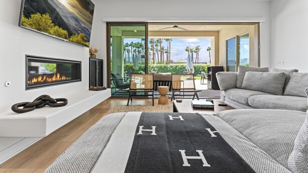 A modern living room with a fireplace, a sofa, and a view of a balcony with palm trees. - The Platinum House!  PGA Signature (Walk to Ernies, Stadium Course!) (La Quinta)