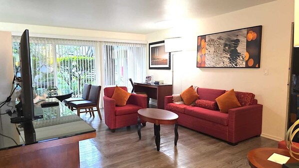 Ein Wohnzimmer mit einem roten Sofa, einem Holztisch, und einem Fernseher. Ein Esstisch mit Stühlen und einer Lampe steht ebenfalls im Raum. Das Zimmer verfügt über ein großes Fenster mit Jalousien.