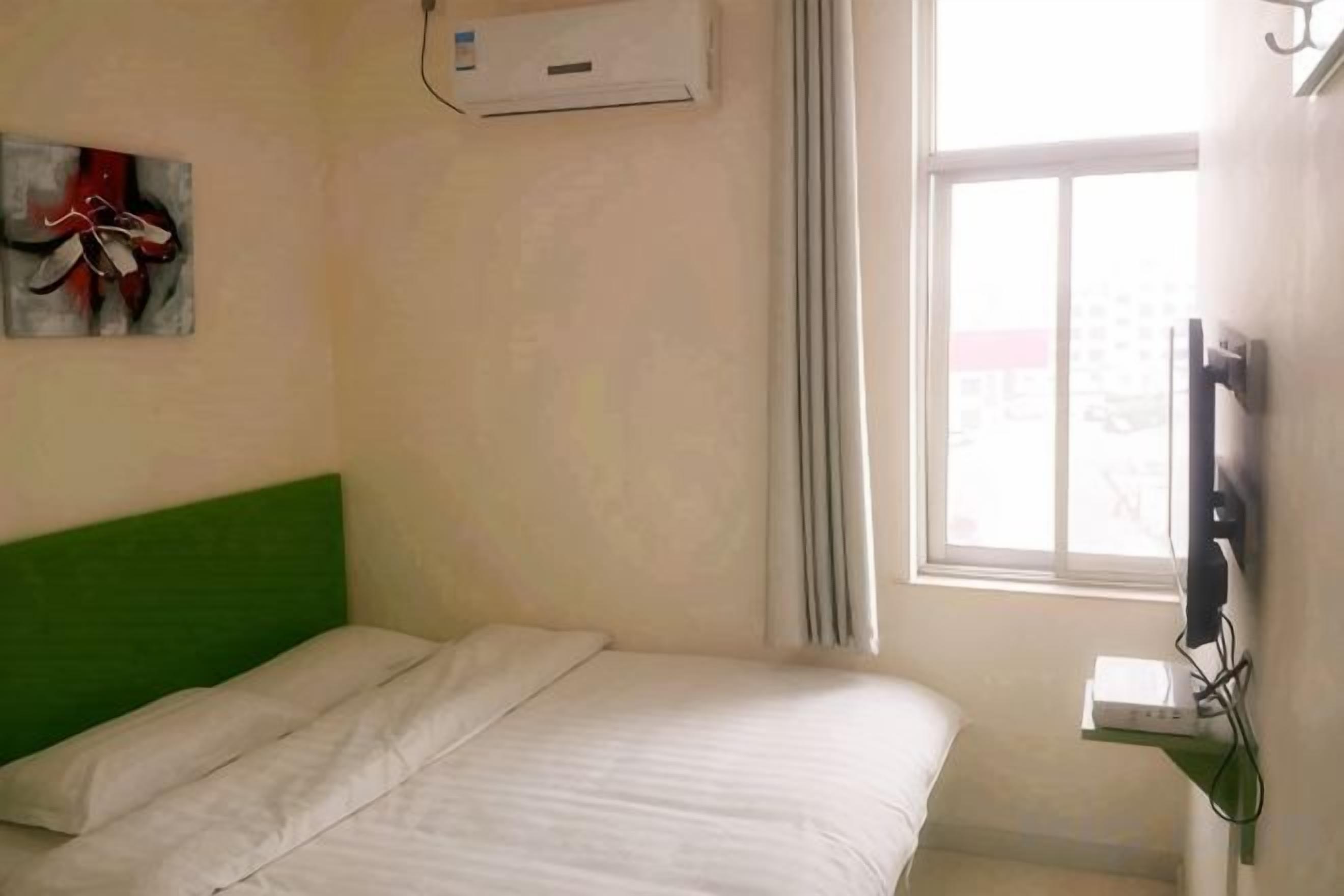 Una cama individual con cabecera verde en una habitación con ventana, equipo de aire acondicionado y televisor de pantalla plana montado en la pared.