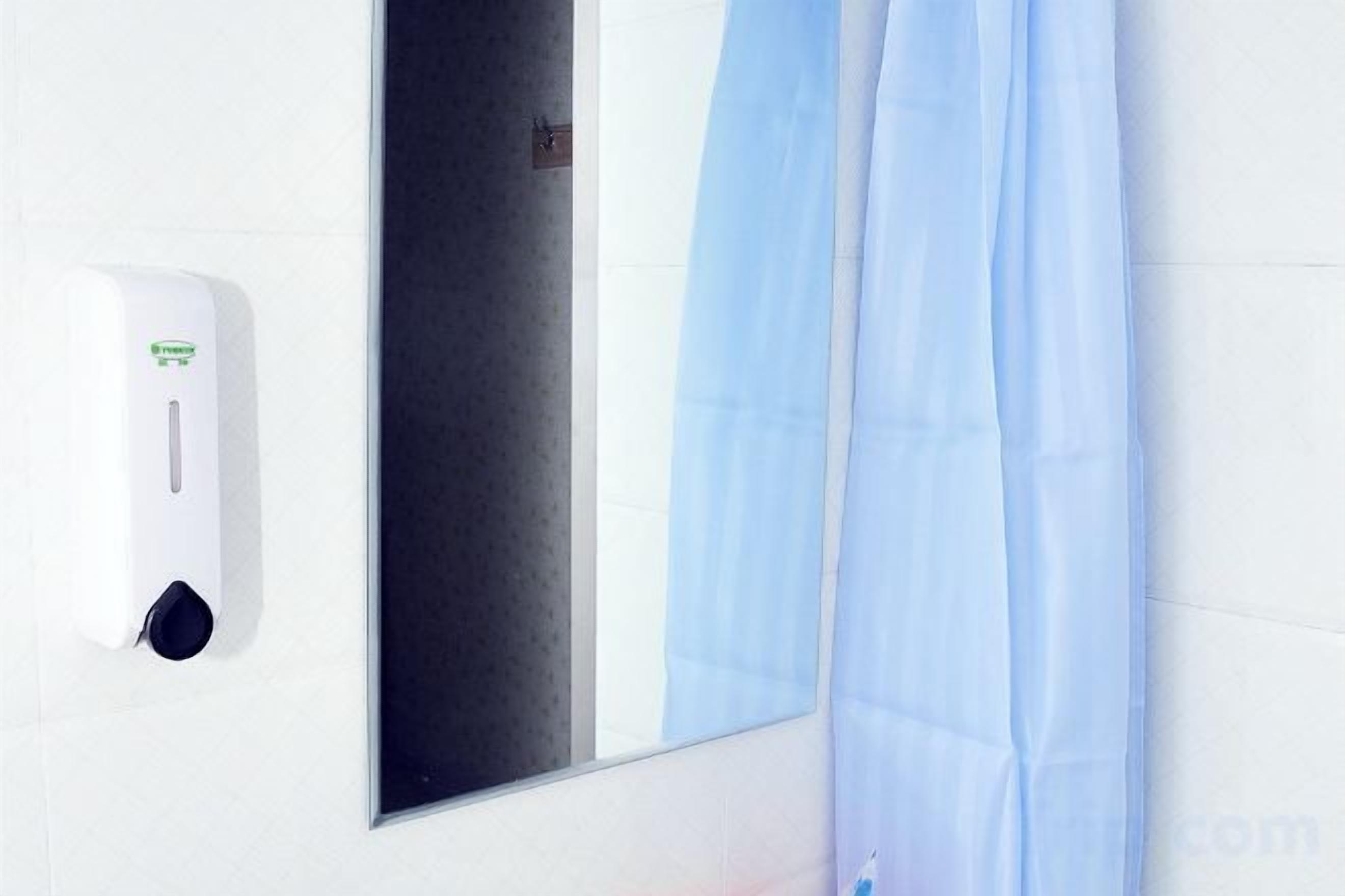 Un distributeur de gel hydroalcoolique fixé au mur, un miroir et un rideau bleu.