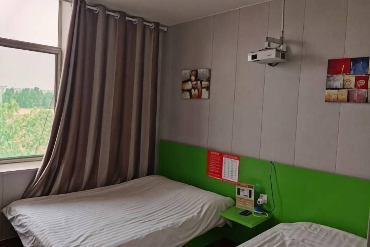 A room with a bed, a green headboard, a window with curtains, a wall-mounted projector, and a bulletin board with notices.
