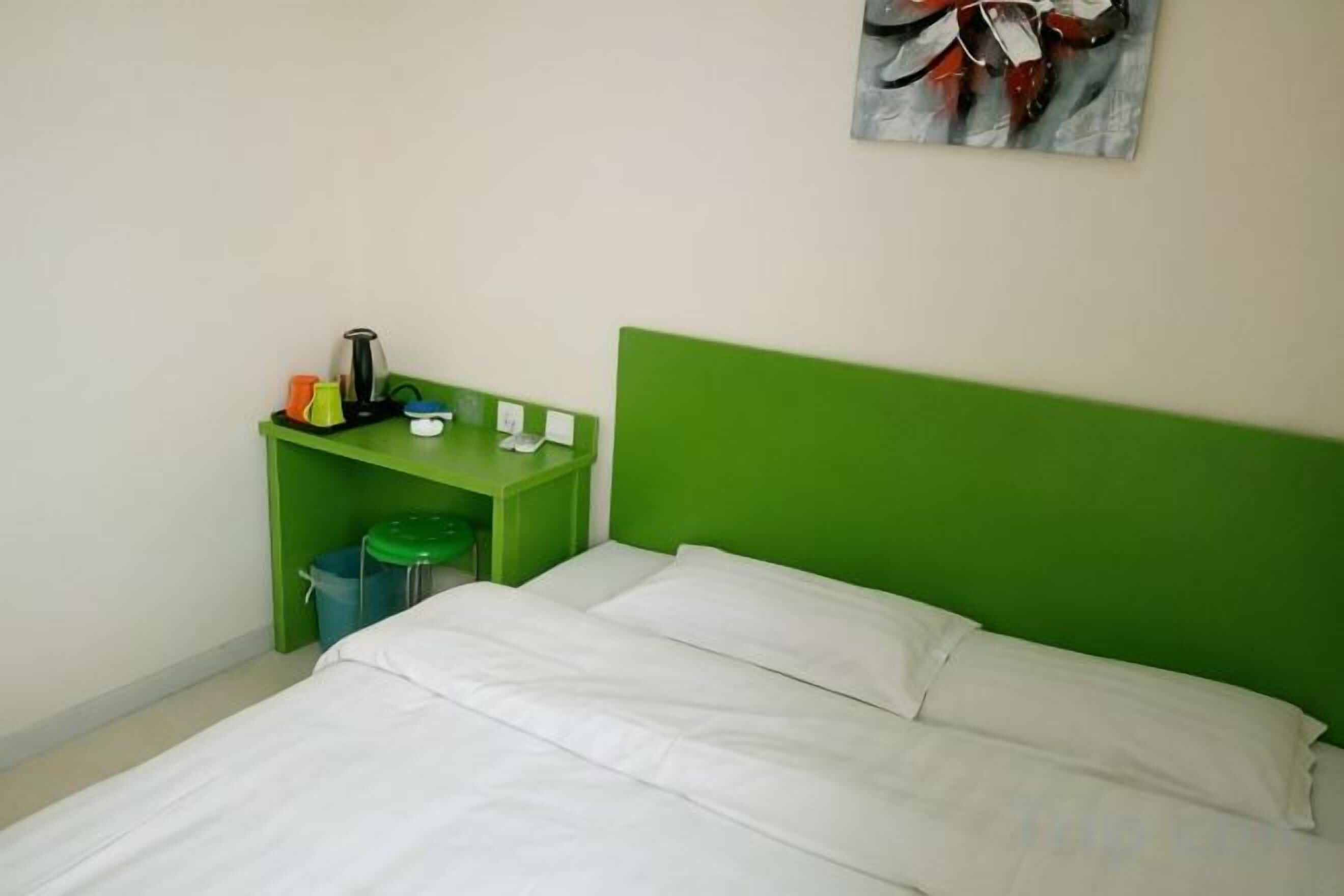 A room with a green headboard, a bed with white bedding, a small table with a kettle, a trash bin, and a wall-mounted light switch.