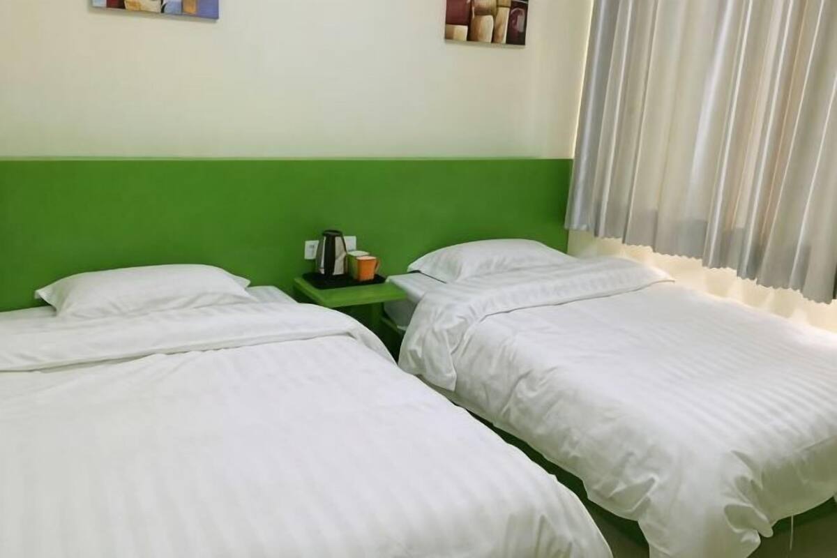 A room with two single beds, a green headboard, a small table with a cup and books, and a window with curtains.