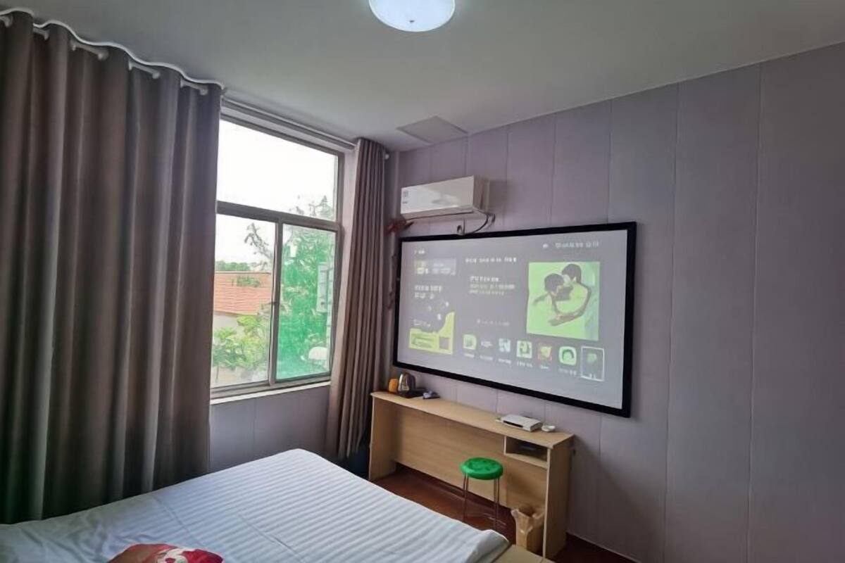 A hotel room with a bed, a desk with a computer, a window with curtains, and a wall-mounted TV displaying a menu.