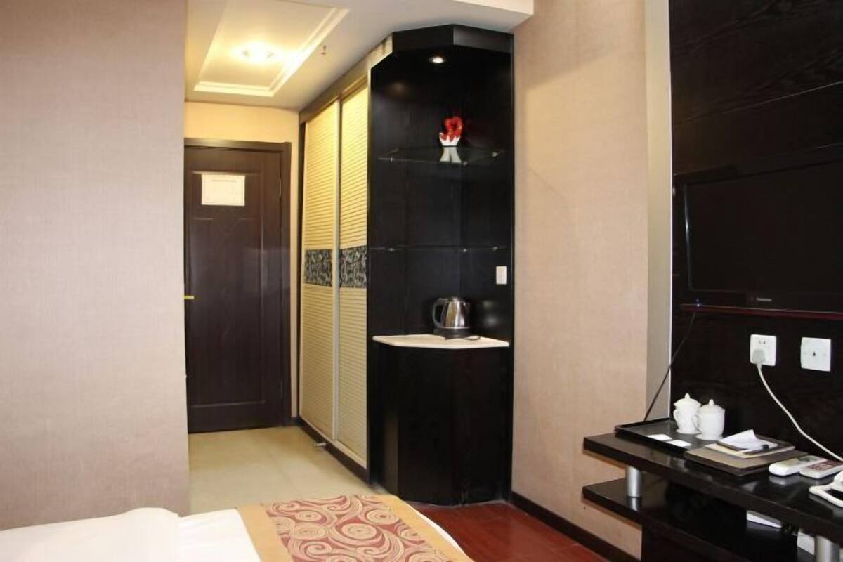 A hotel room with a bed, a small bedside table with a lamp, a built-in shelf with a vase, and a wall-mounted TV.